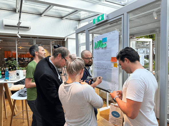 Das Bild zeigt Teamarbeit im Mob, Mobbing. Mehrere Menschen stehen um einen Flipchart und schreiben gemeinschaftlich an einer fiktiven E-Mail. Alle wirken sehr konzentriert.
