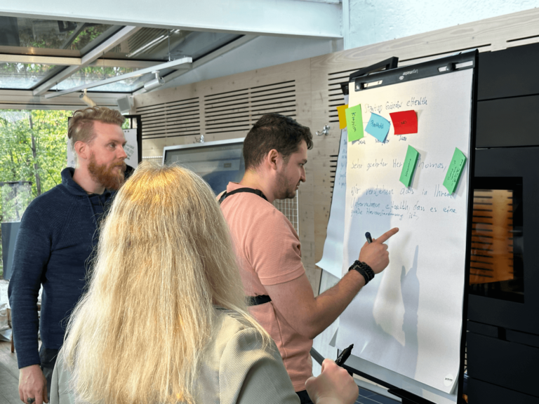 Das Bild zeigt Teamarbeit im Mob, Mobbing. Mehrere Menschen stehen um einen Flipchart und schreiben gemeinschaftlich an einer fiktiven E-Mail. Alle wirken sehr konzentriert.