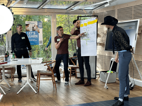 Das Ergebnis der Teamarbeit (Mobbing) wird von einem Mobmitglied vorgestellt. Der Mann hält ein Mikrofon in der Hand und deuetet mit der linken Hand auf das Flipchart.